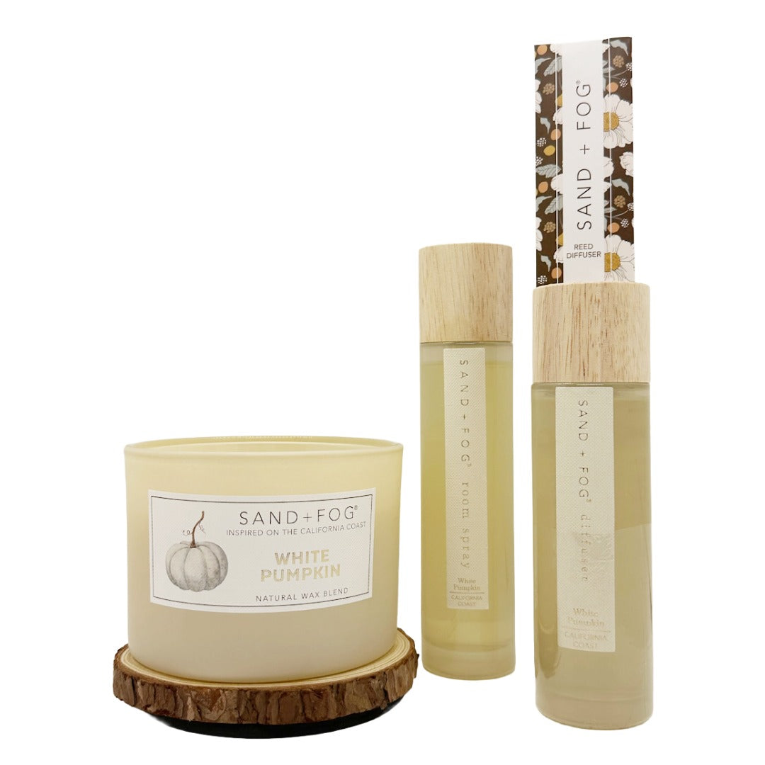 A Sand + Fog natural wax candle in a cream-coloured jar with a wood slice coaster, shown alongside matching room spray and reed diffuser bottles with light wood caps on a white background.