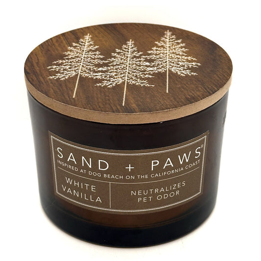 A dark amber glass jar candle with a round wooden lid featuring an engraved white pine tree design. The front label reads 'SAND + PAWS' and 'WHITE VANILLA'.