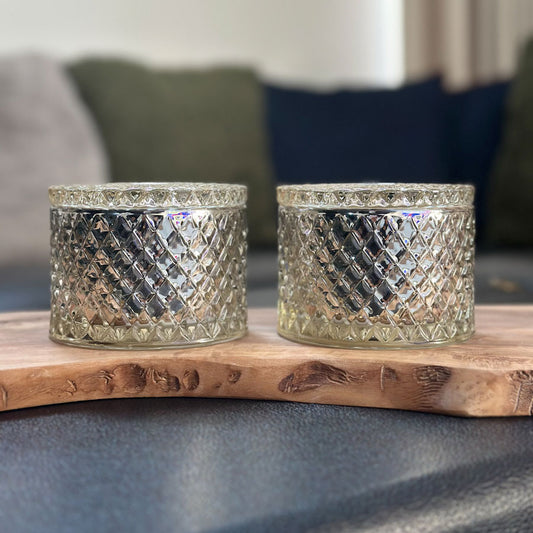 Two winter pine decorative glass candles on a wooden surface with a blurred background.